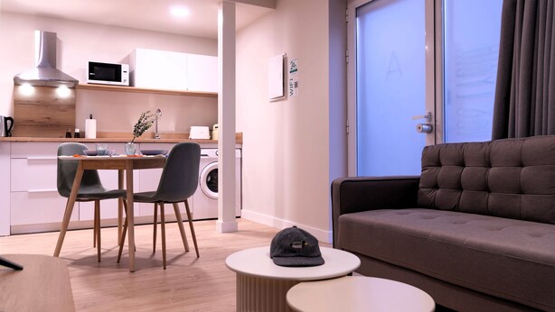 A modern living room with a sofa and a dining area with a table and chairs. There is a washing machine and a microwave in the kitchen area.