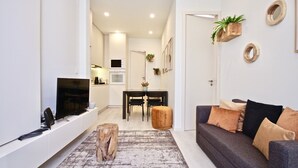 A modern living room with a grey sofa, a coffee table, and a TV mounted on the wall. There is a dining area with a table and chairs, and a kitchen in the background. - Marjoram Apartment, Santos, Lisbon (Lisboa)