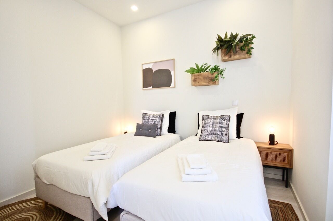 A hotel room with two single beds, white bedding, a small bedside table with a lamp, a wall-mounted plant, and a framed abstract artwork.