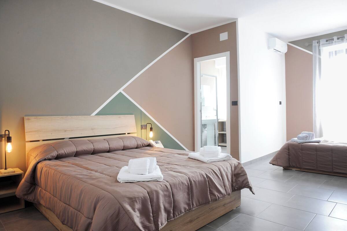 A modern hotel room with a large bed, a bedside table with a lamp, a mirror, and a view of the hallway.