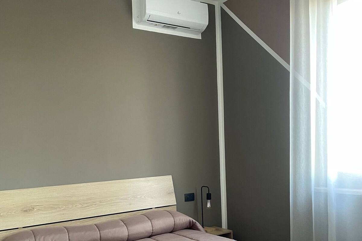 A bedroom with a bed, an air conditioning unit, a nightstand, and a window with sheer curtains.
