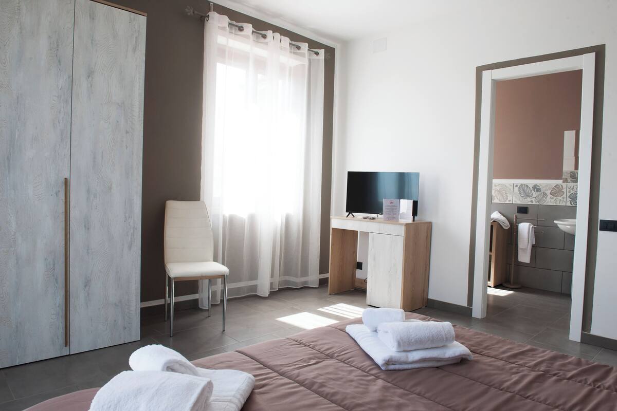 A modern hotel room with a bed, a desk with a TV, a chair, and a bathroom visible through an open door.