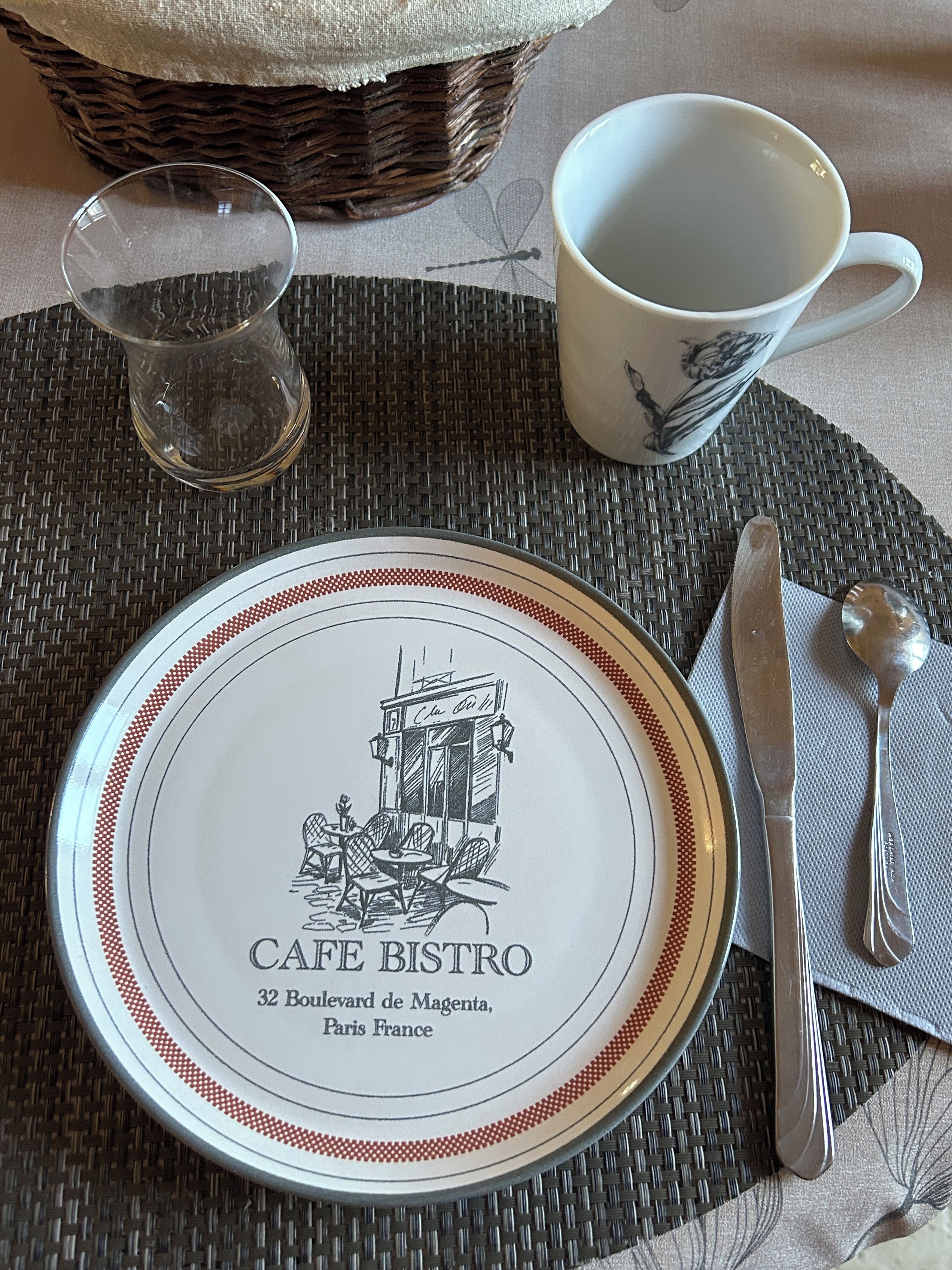 A dining setup with a plate labeled 'Café Bistro,' a cup, a glass, a knife, and a spoon on a woven placemat.