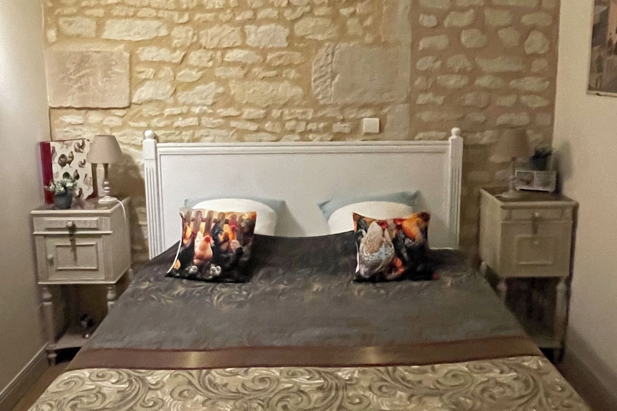 A bedroom with a stone wall, a bed with patterned bedding, a nightstand with a lamp, and a small table with a picture frame.