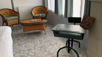 Una habitación con baldosas de diseño, una mesa verde, dos sillas de mimbre, un radiador y una ventana con cortinas.