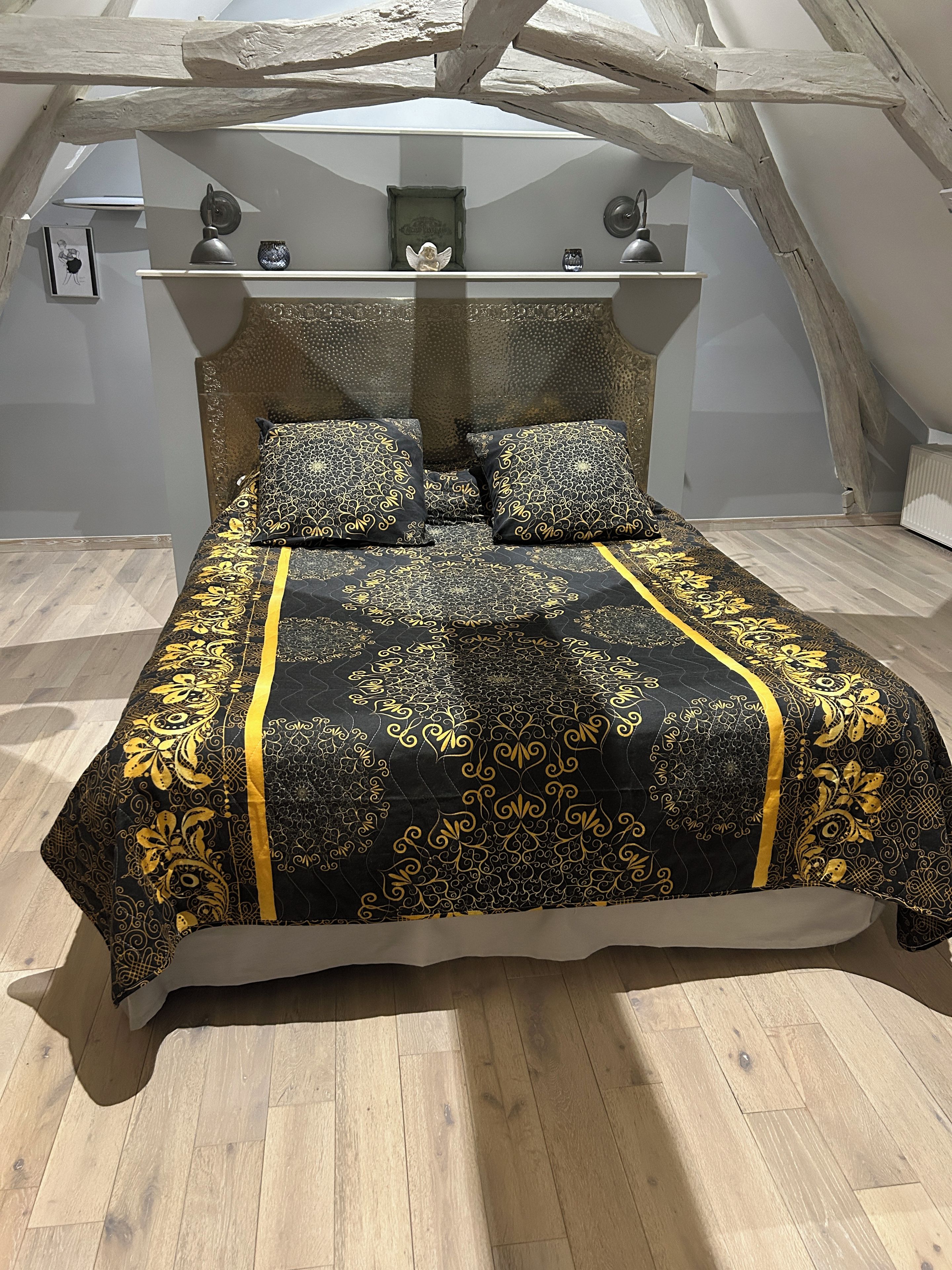 Ein Bett mit einer schwarz-goldenen Bettdecke und passenden Kissen, ein hölzernes Kopfteil und eine weiße Wand mit einem kleinen Bilderrahmen und Dekorationselementen.
