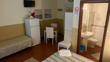 Ein kleines Hotelzimmer mit einem Bett, einem Schreibtisch, einem Stuhl, einem Kühlschrank, einem Fernseher und einem Badezimmer mit Waschbecken und Toilette.