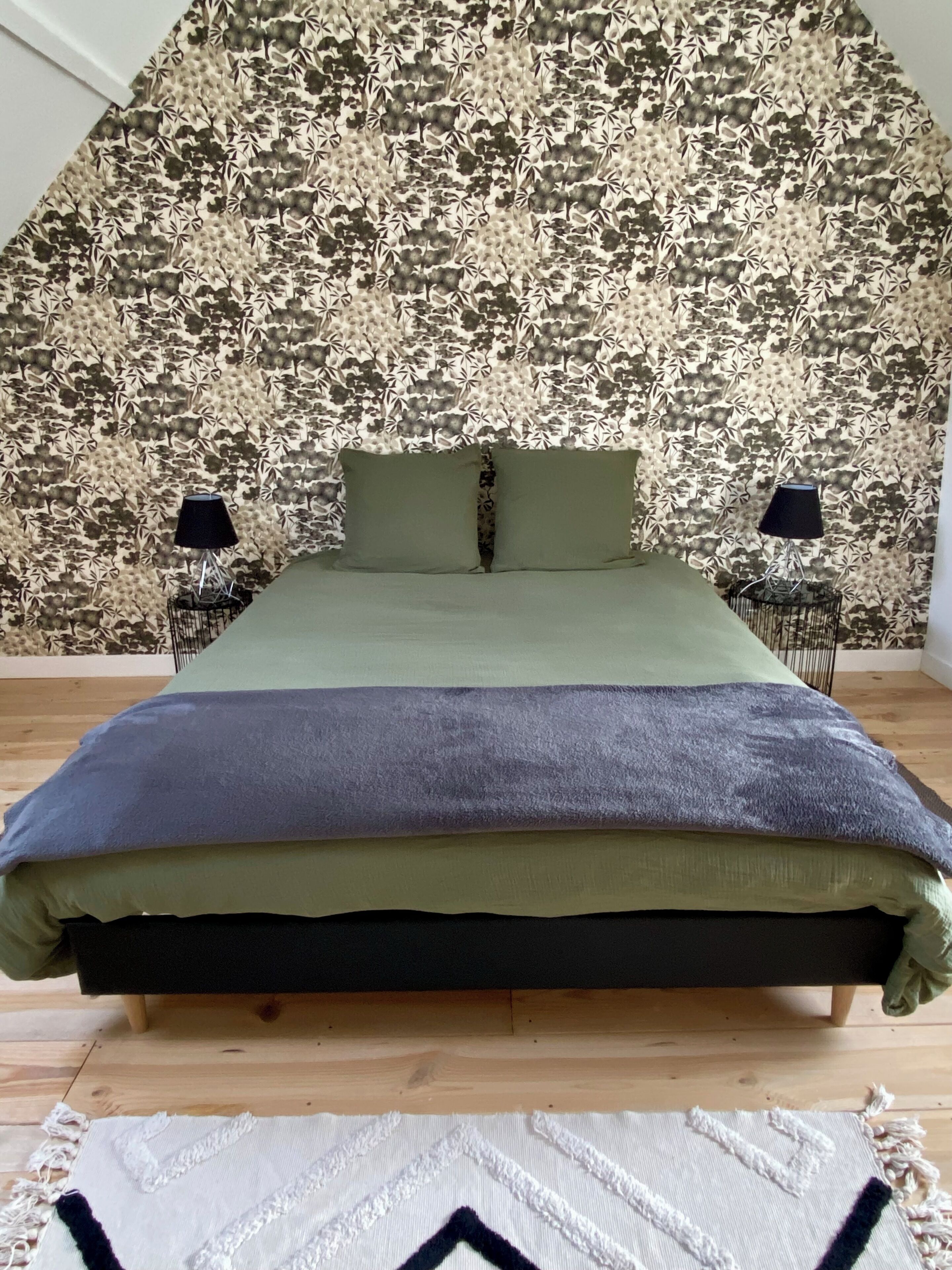 A bed with a green and purple bedspread, two pillows, and a floral wallpaper background. There are two bedside tables with lamps.