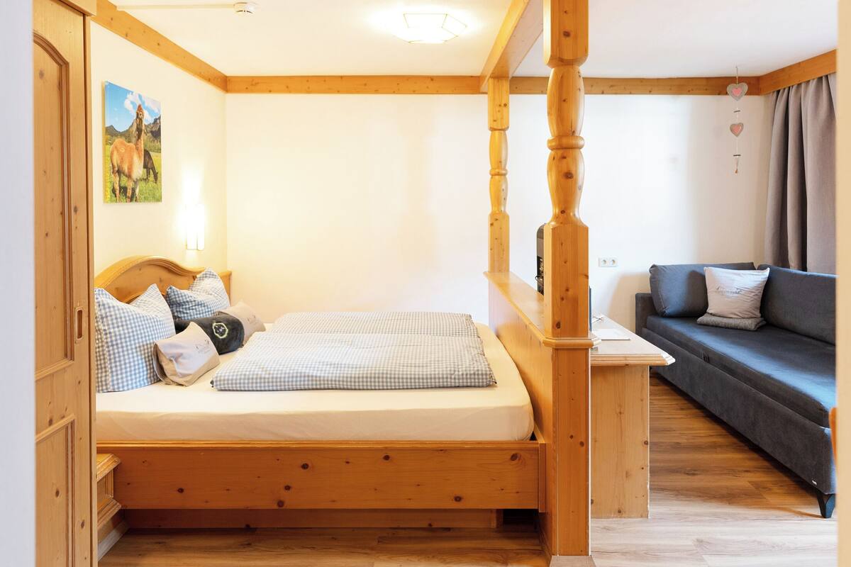 Een slaapkamer met een houten bed, een bank en een bureau. Er hangt een foto aan de muur en er is een raam met gordijnen.