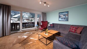 A living room with a grey sofa, a red chair, a wooden coffee table, and a large window with a view of a snowy mountain and buildings. - Käserstube Ferienwohnungen (Tannheim)