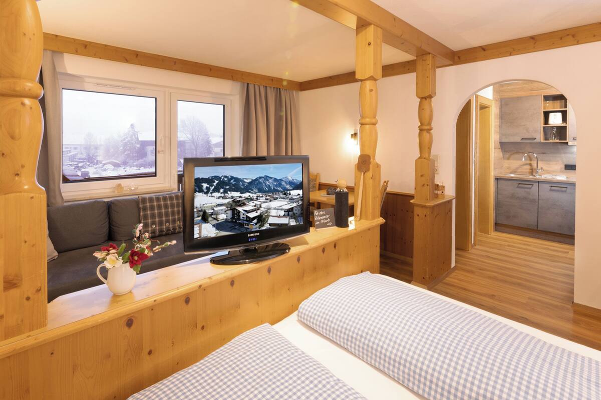 Een gezellige kamer met een houten hoofdeinde, een televisie op een houten standaard, een bank, een bed met geruit beddengoed en uitzicht op een besneeuwde stad door het raam.