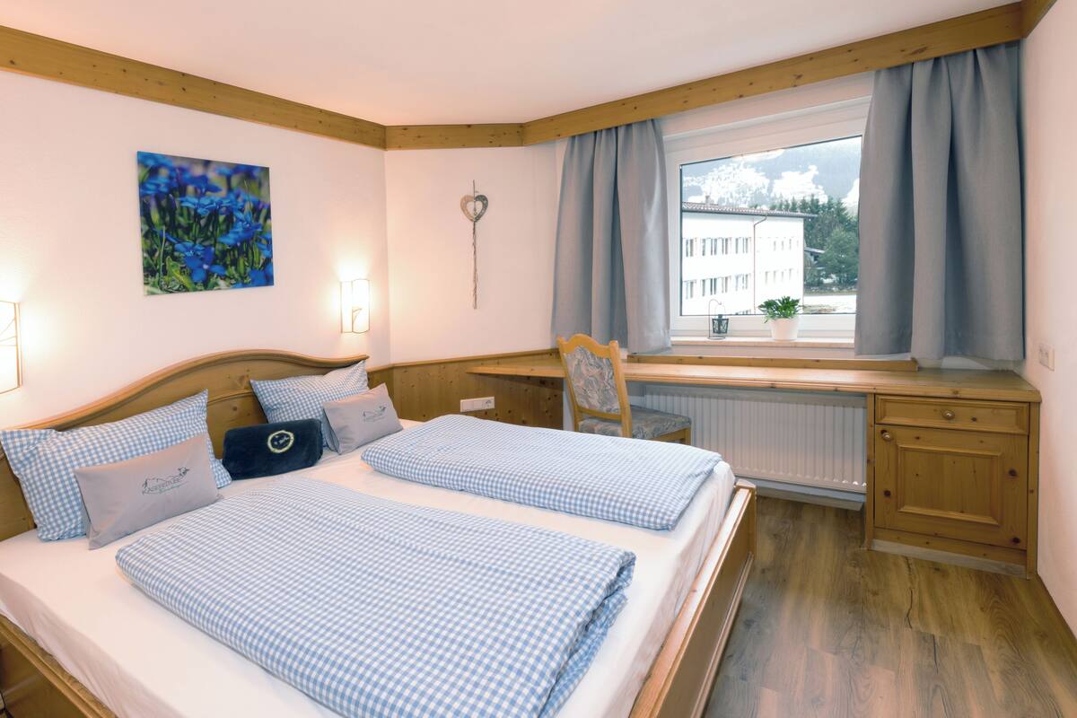 Een tweepersoonsbed met geruit beddengoed, een nachtkastje, een stoel, een raam met gordijnen, en een schilderij aan de muur.