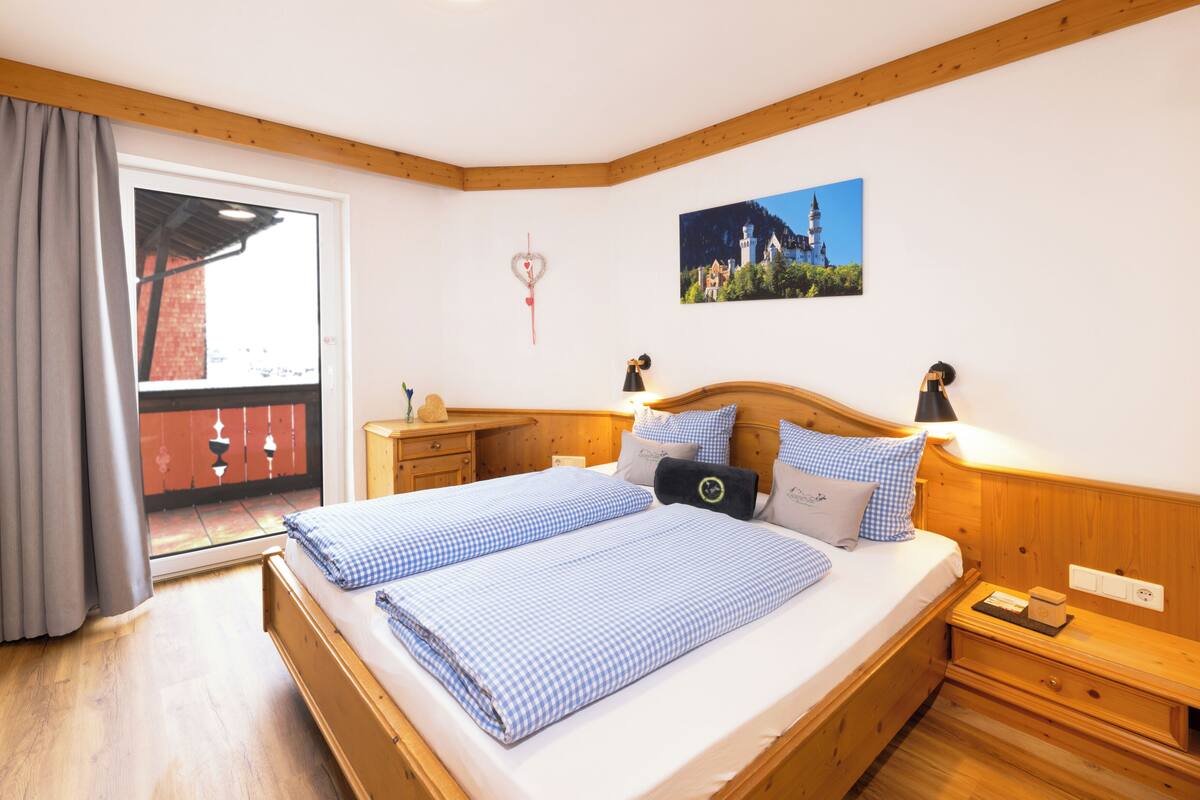 Een slaapkamer met een houten bed, een nachtkastje, een dressoir, een raam met gordijnen en een foto aan de muur.