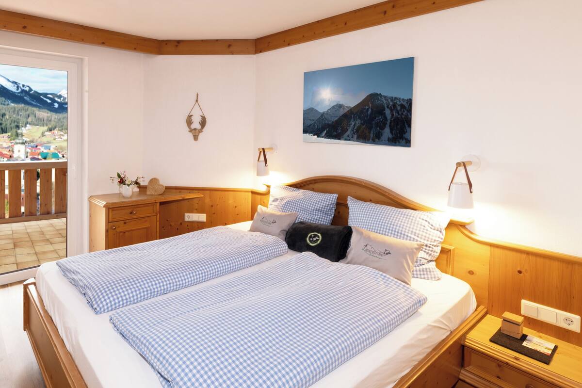 Een slaapkamer met een groot bed, een houten hoofdbord, nachtkastjes, een nachtkast en een raam met uitzicht op bergen, plus een wandlamp.