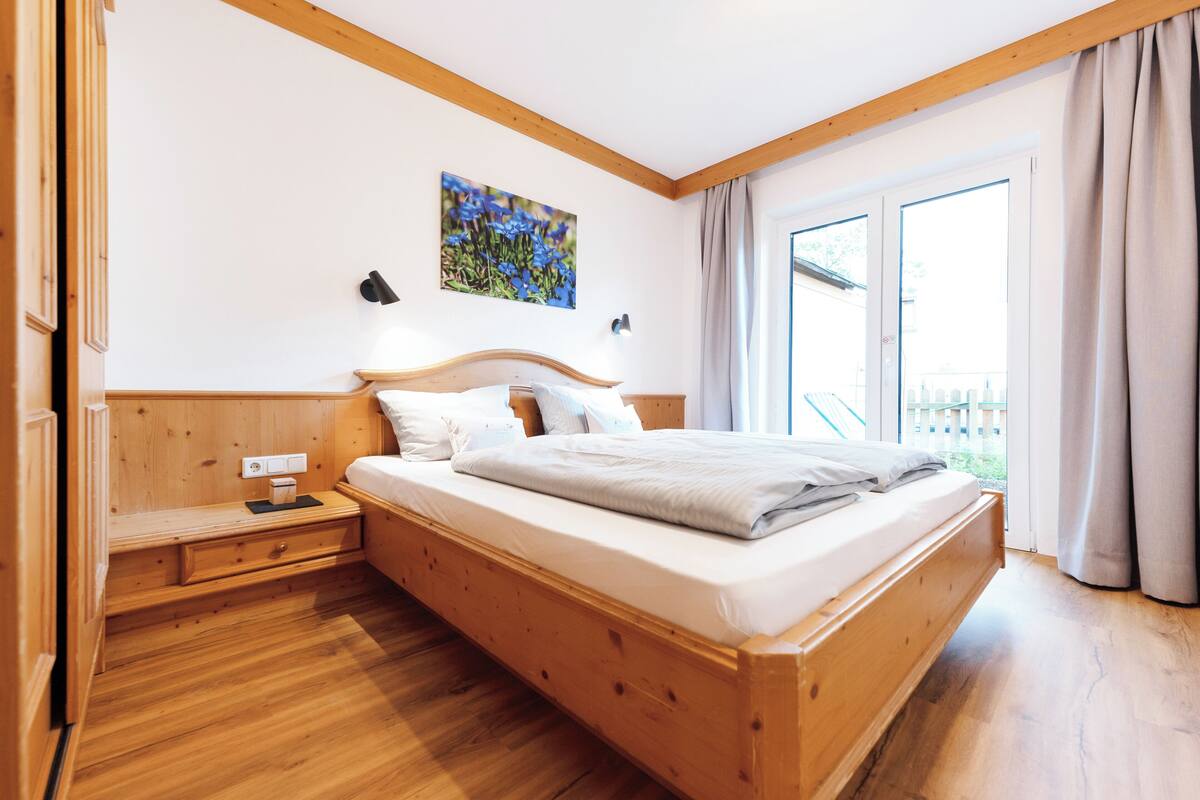 Een houten bed met hoofdeinde, een nachtkastje en een wandlamp. Er is een raam met gordijnen, en een schilderij boven het bed.