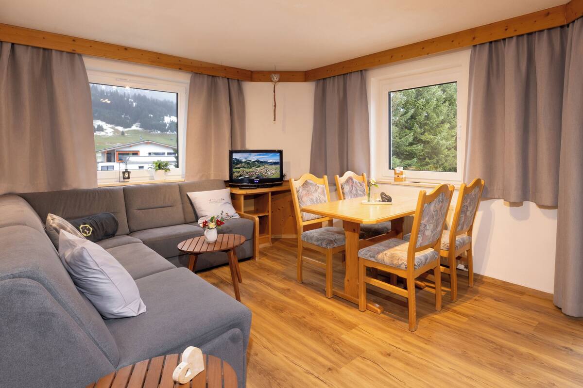 Een gezellige woonkamer met een bank, een klein rond tafeltje, een houten eettafel met stoelen, een televisie en grote ramen met gordijnen.