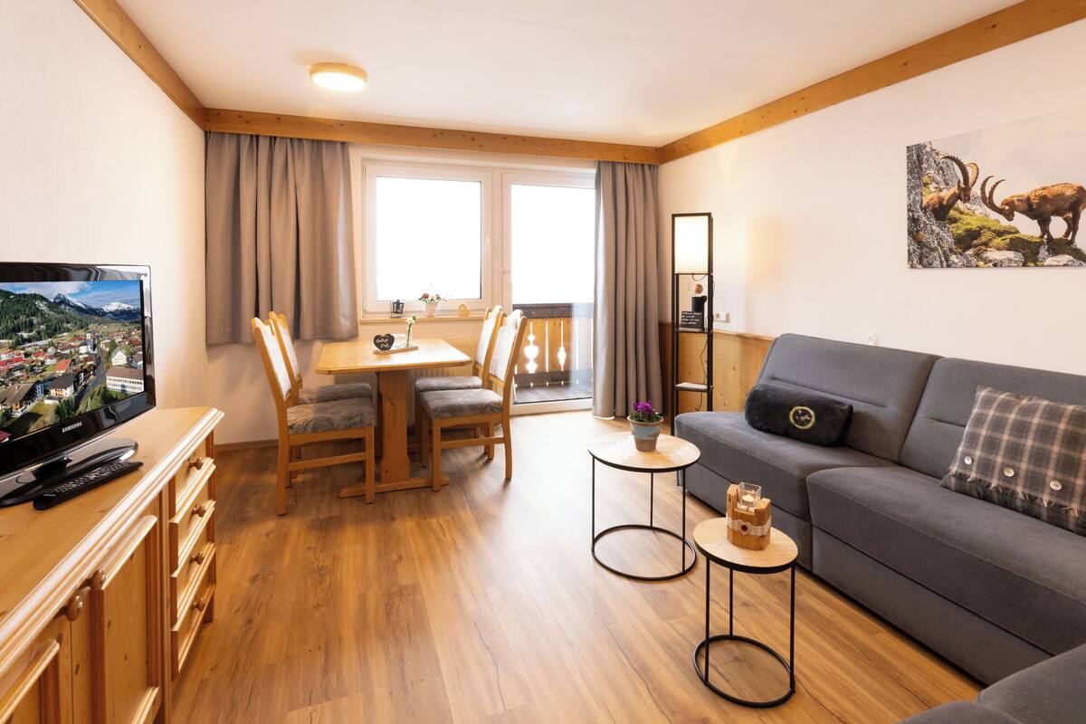 Een gezellige woonkamer met een grijze bank, een houten eettafel en stoelen. Een flatscreen televisie staat op een houten kast, en er is een klein tafeltje met een kaars en een decoratief object.
