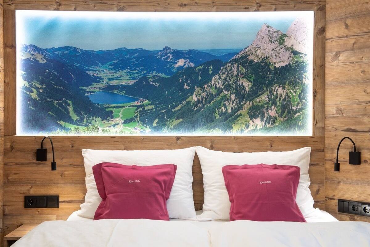 Een slaapkamer met een groot schilderij van een berglandschap boven het bed, twee kussens met een merknaam, een houten hoofdbord en een nachtkastje met een lamp.