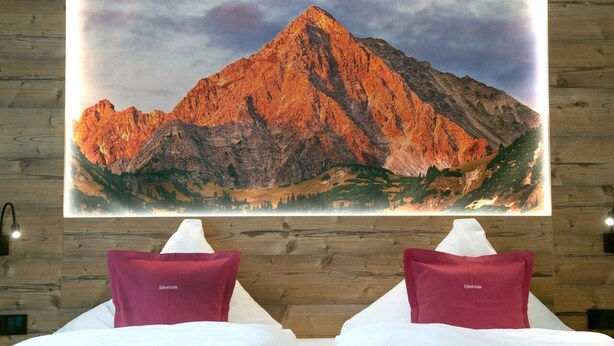 A hotel room with a large mountain painting on the wall, two red pillows, and a wooden headboard. - Käserstube Ferienwohnungen (Tannheim)