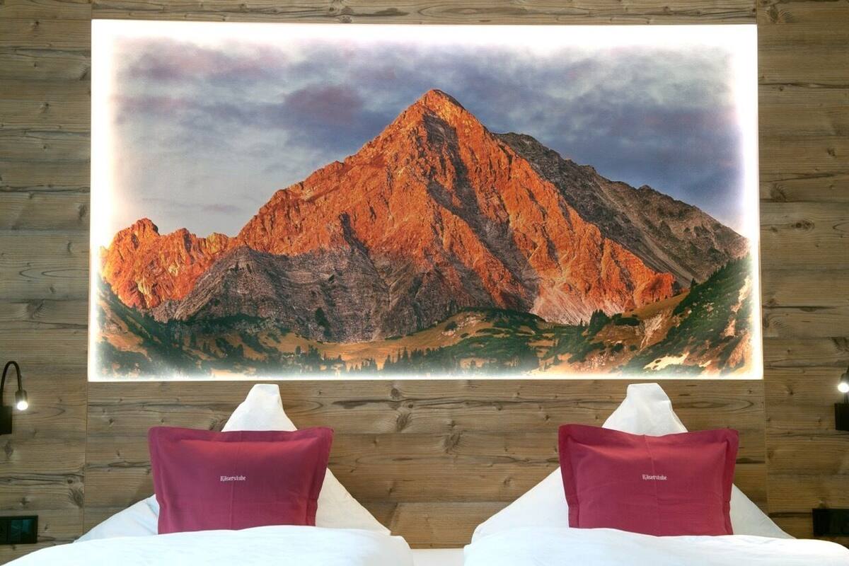 Een hotelkamer met een groot berglandschap op de muur, twee rode kussens en een houten hoofdbord.