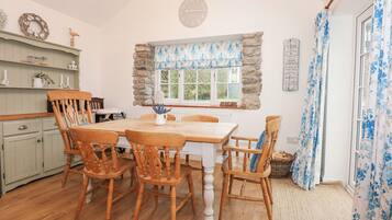 A cozy kitchen with a wooden table and chairs, a stone accent wall, a window with blue floral curtains, and a clock on the wall.