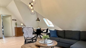 A modern loft apartment with a high ceiling, a dark grey sectional sofa, two round wooden tables, a black wicker chair with a fur throw, and a skylight. - Apartment in Sint Maartenszee Near Beach (Sint Maartensvlotbrug)