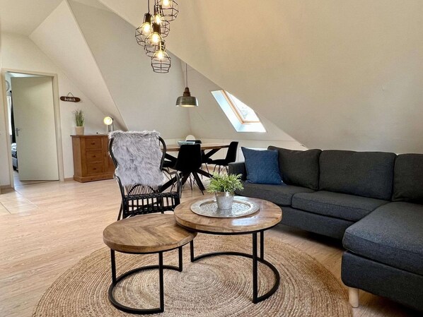 A modern loft apartment with a high ceiling, a dark grey sectional sofa, two round wooden tables, a black wicker chair with a fur throw, and a skylight.