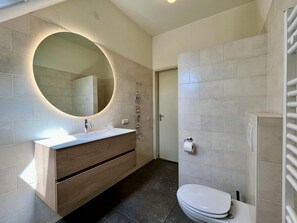 A modern bathroom with a round mirror, wooden vanity, white toilet, and tiled walls.