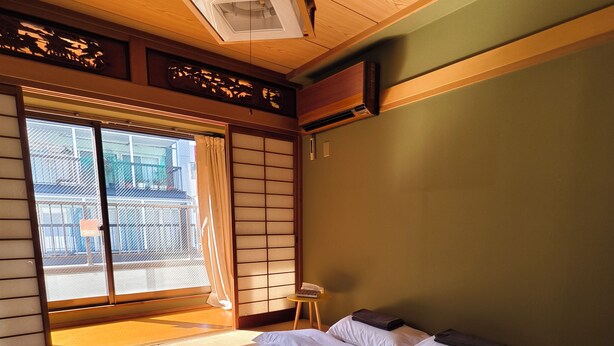 Um quarto em estilo japonês tradicional com tatames, uma cama grande com roupa de cama branca, portas deslizantes e vista para um edifício lá fora.