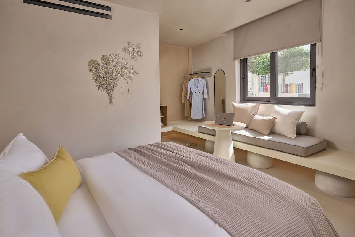 A modern bedroom with a bed, a bench with pillows, a laptop on a small table, and a window with a view of the outside.