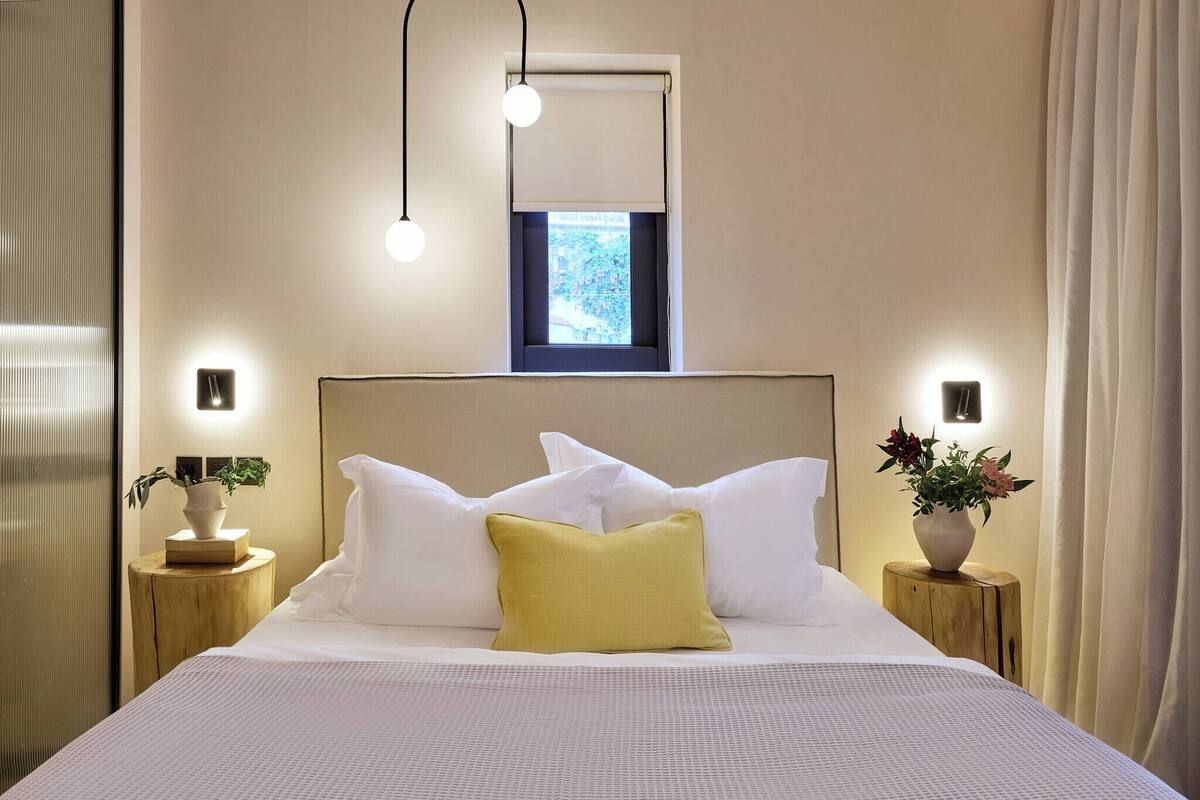 A neatly made bed with white and yellow pillows, a small window with a view, and bedside tables with vases.