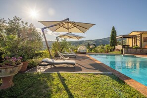 A poolside area with lounge chairs and umbrellas, surrounded by greenery and a view of hills. - Villa Alessia Pool Wellness And Nature  (Massa e Cozzile)