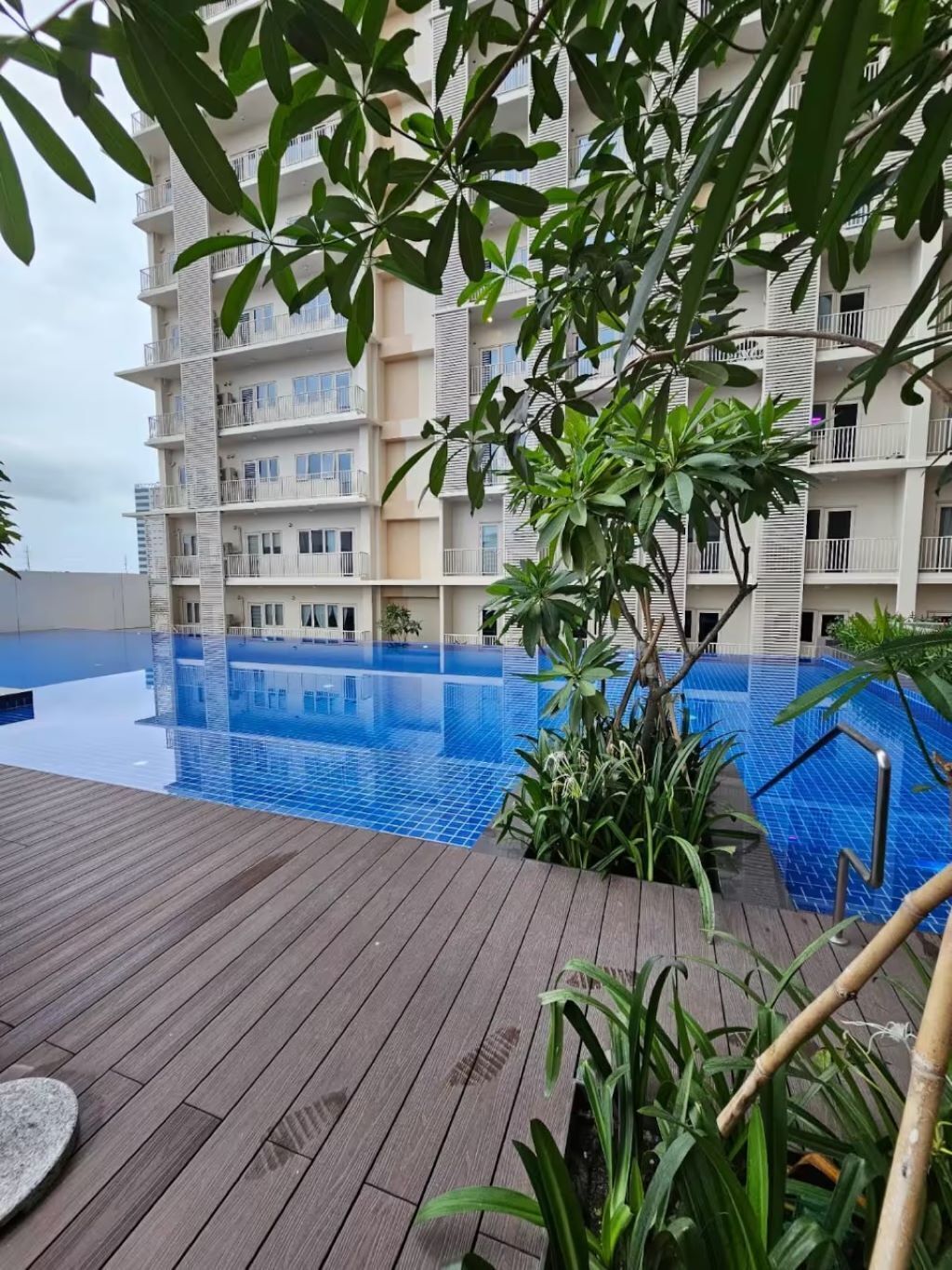 A rooftop pool area with a wooden deck, surrounded by lush greenery and a multi-story building with balconies.