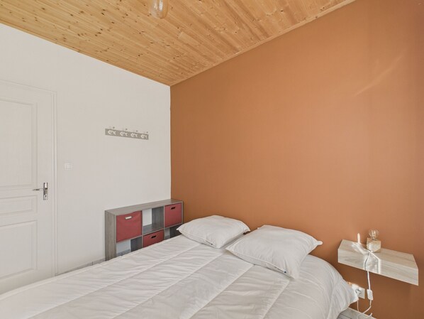 A bedroom with a wooden ceiling, a white door, a bed with white bedding, a nightstand with a lamp, and a wall-mounted shelf.