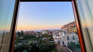 Vista a través de una ventana de un pueblo costero con edificios, vegetación y una colina al fondo.
