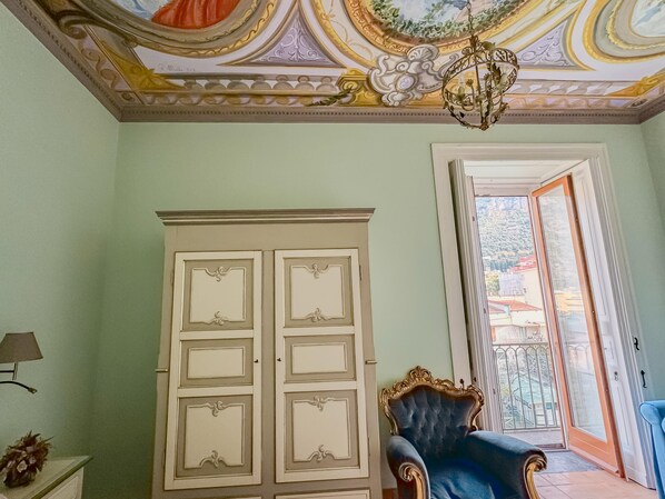 A room with a large white wardrobe, a blue upholstered chair, a chandelier, and a view through a door to a balcony with a railing. - Vanvitelli Palace B&b (Meta)