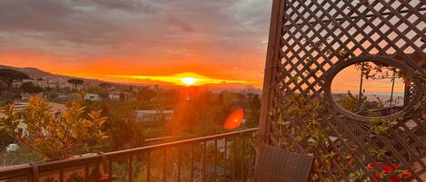 A balcony with a glass table and wicker chairs, overlooking a sunset with buildings in the distance.