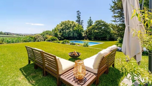 A patio with wooden furniture and a table, a small fire pit, and a pool in the background.