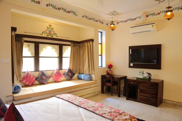 Un dormitorio con cama, un asiento junto a la ventana con cojines, escritorio con silla, televisor y un mueble pequeño.