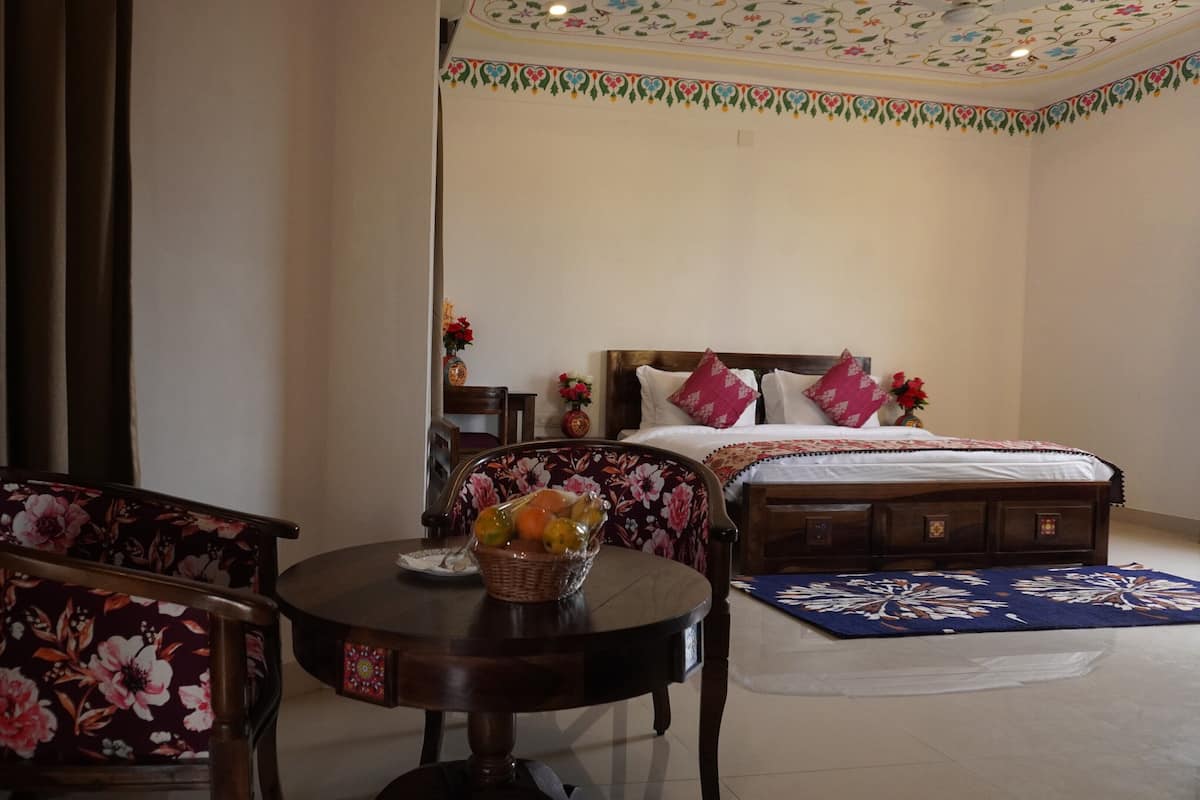 Una habitación de hotel con una cama, dos sillas, una mesa con una cesta de frutas y un techo decorativo.