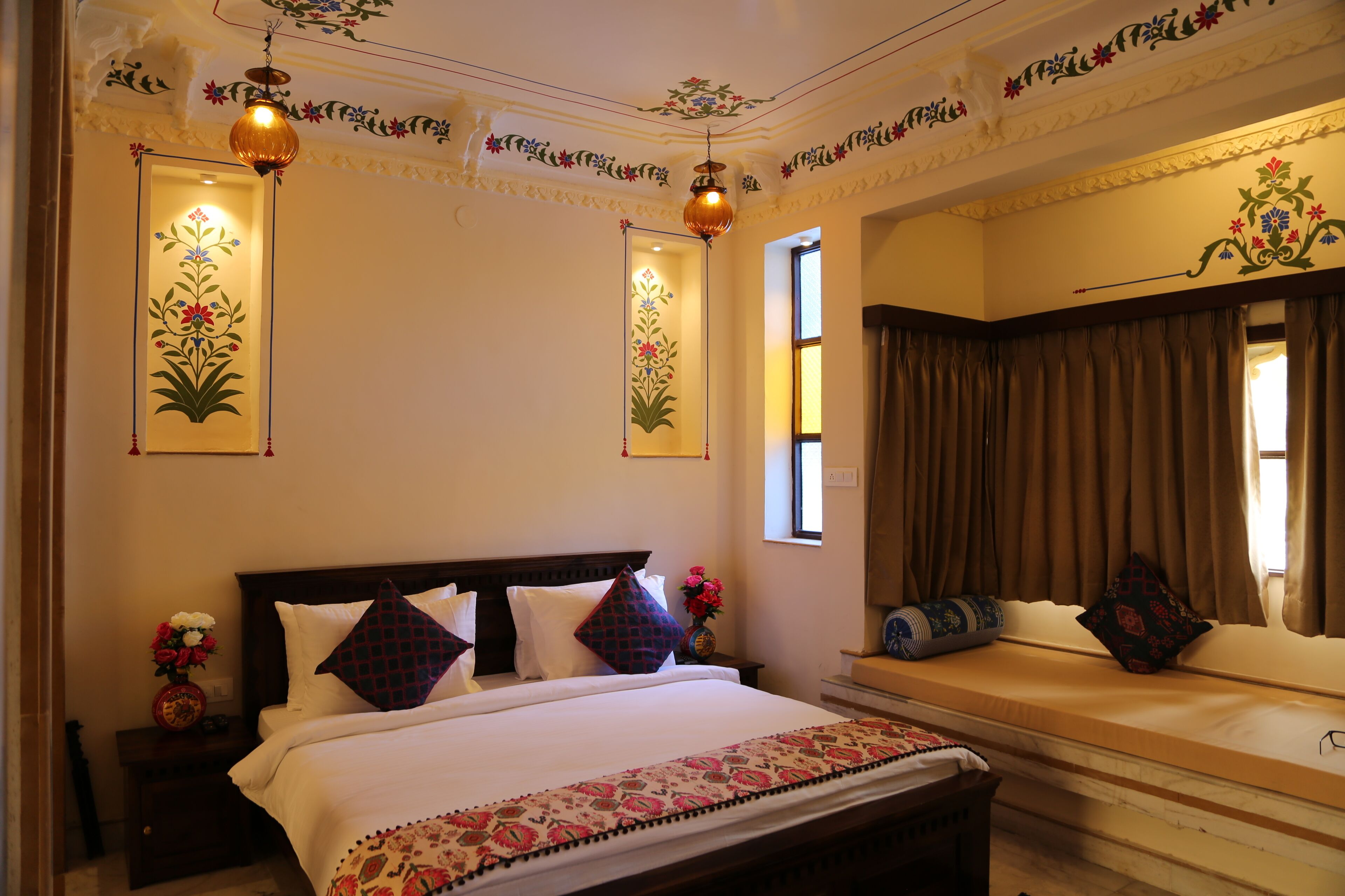 A well-lit bedroom with a large bed, a bedside table with a vase of flowers, and a window with curtains.