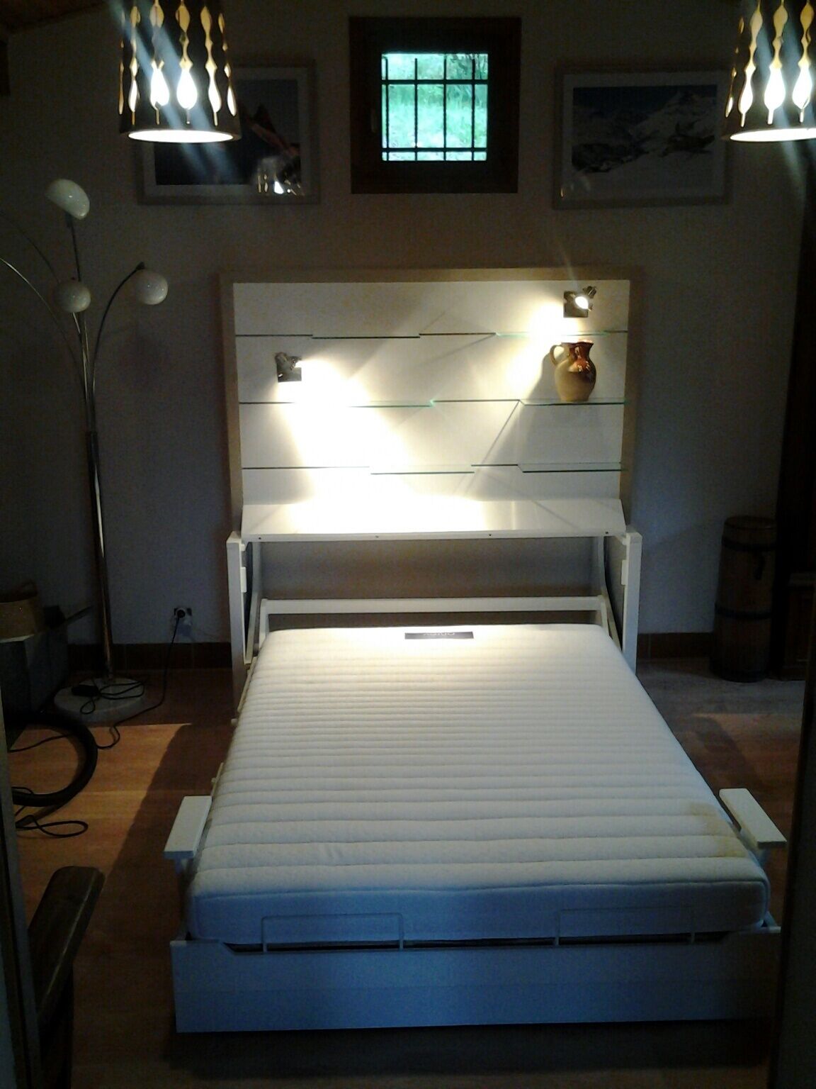 Une chambre à coucher avec un lit, une tête de lit dotée de lampes, une fenêtre et une étagère fixée au mur, sur laquelle se trouve un vase.