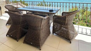 A terrace with wicker furniture, a table, and a view of the sea and distant hills.