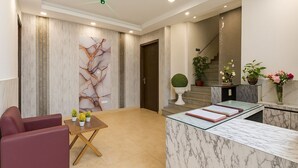 A modern reception area with a marble reception desk, a seating area with a table and chairs, and a staircase leading to an upper floor. - Hotel Rajvansh Palace (Gurugram)