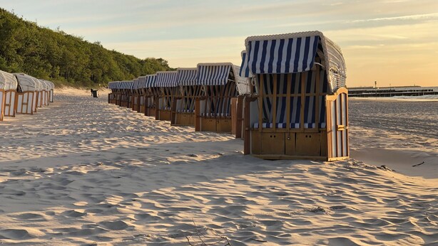 A row of beach huts with striped awnings, wooden frames, and cushioned seating, lined up along a sandy beach with a forested hill in the background.