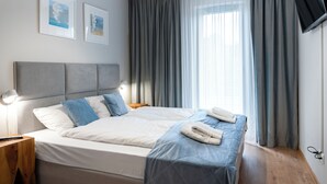 A double bed with a grey headboard, two pillows, a blue blanket, and a folded towel. There is a bedside table with a lamp, a TV mounted on the wall, and two framed pictures on the wall.