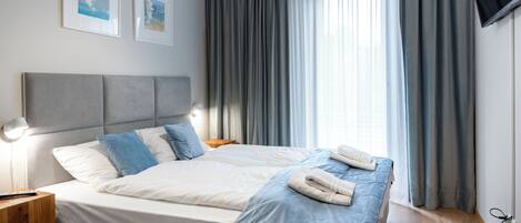 A double bed with a grey headboard, two pillows, a blue blanket, and a folded towel. There is a bedside table with a lamp, a TV mounted on the wall, and two framed pictures on the wall.