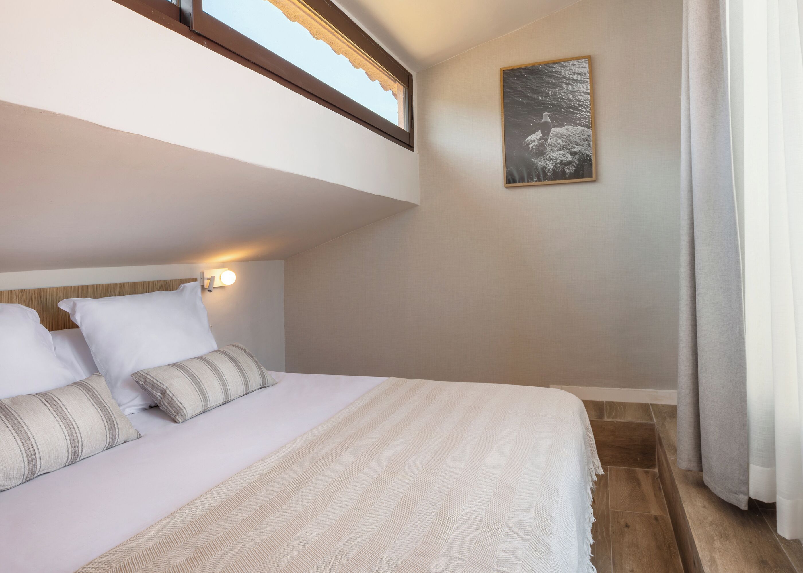 Un dormitorio con cama, almohadas, cabecero, luz empotrada en la pared, un cuadro enmarcado y una ventana.