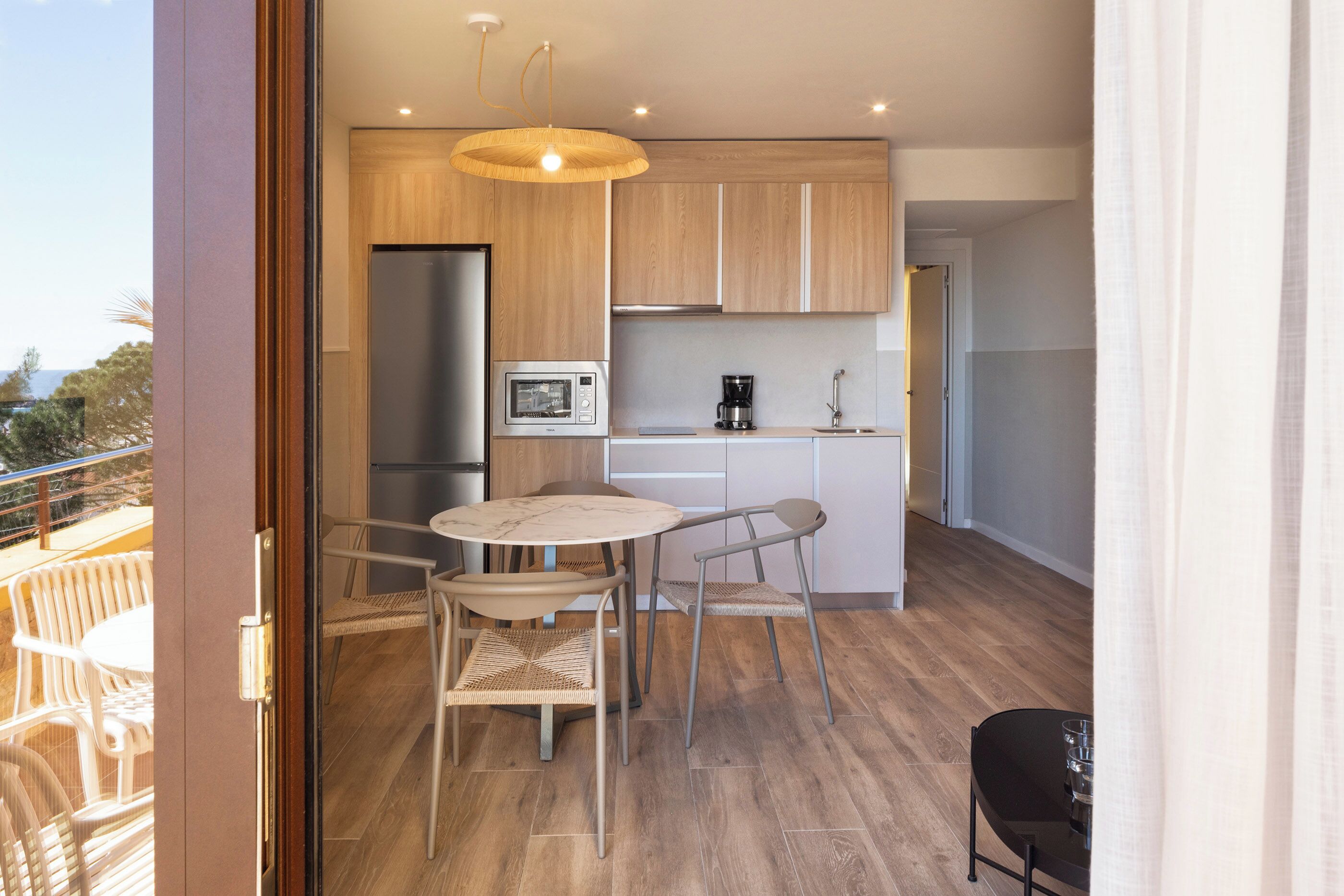 A modern kitchen with wooden cabinets, a stainless steel refrigerator, a microwave, and a coffee maker. There is a dining area with a round table and two chairs.