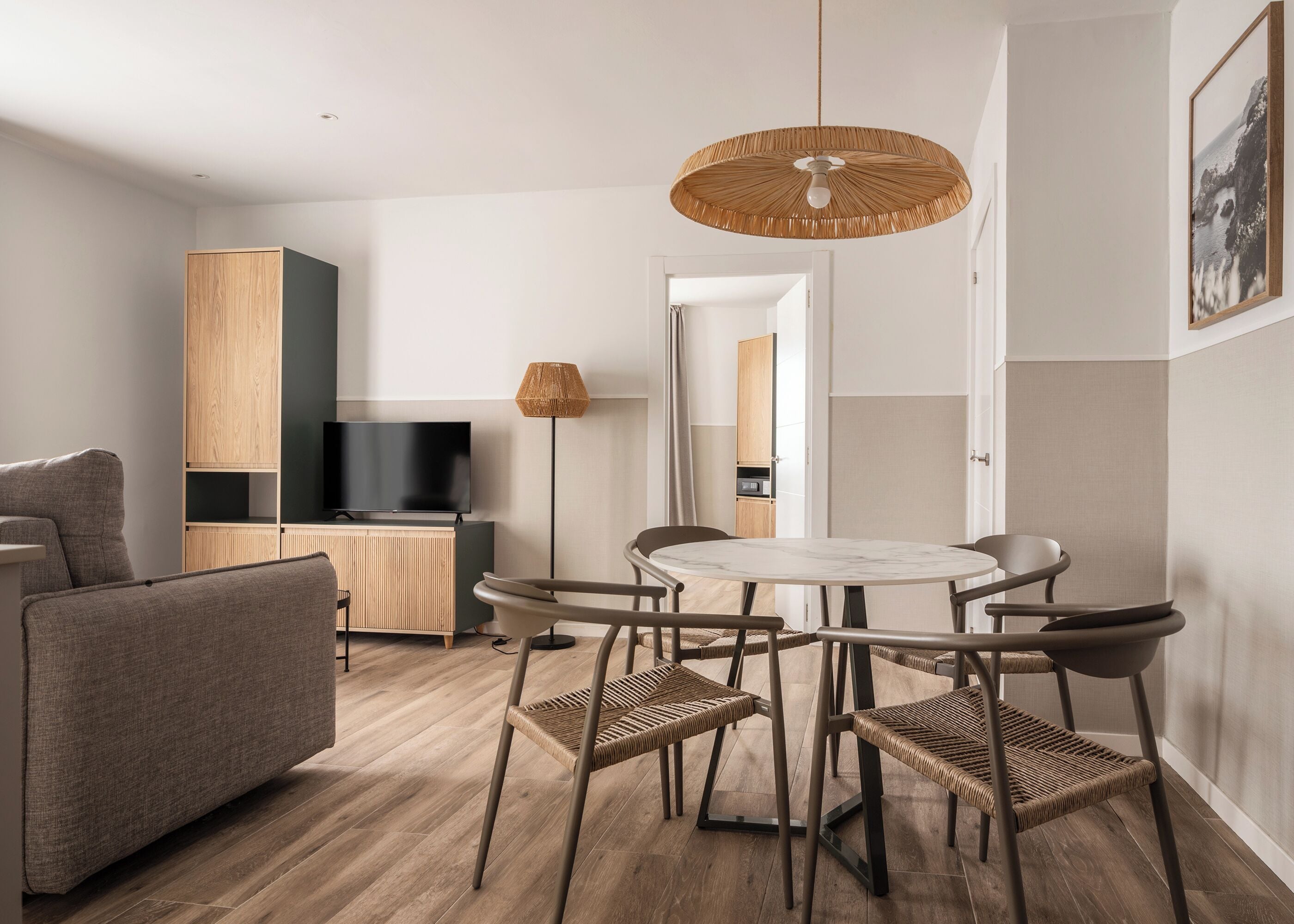 A modern living room with a round dining table, wicker chairs, a flat-screen TV, and a woven ceiling light fixture.